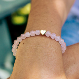 Pink rose quartz beaded bracelet with a gold heart charm on a wrist against a blurred background