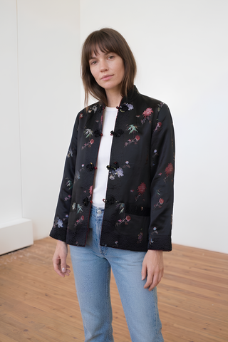 Woman wearing a black floral jacket over a white shirt and blue jeans in a minimal indoor setting.
