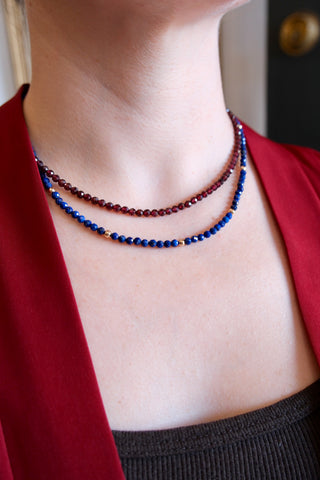 Close up of model wearing a faceted garnet beaded necklace and a faceted blue lapis and gold bead necklace stacked.