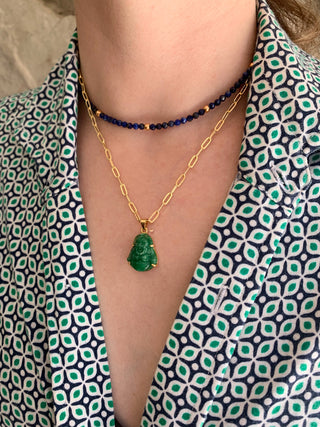 Close up of a person wearing a gold necklace with a jade Buddha pendant and a blue lapis necklace, against a patterned garment.