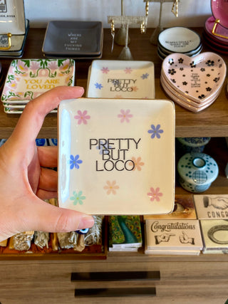 Hand holding a small square dish with 'Pretty But Loco' text against a background of decorative items.