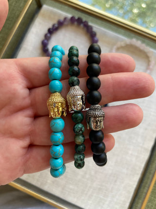 Three beaded bracelets with Buddha charms held in a hand, with a blurred background.