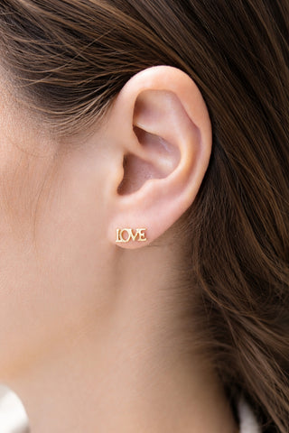 A close-up of a person's ear wearing a small gold stud earring with the word 'LOVE' spelled out in tiny letters.