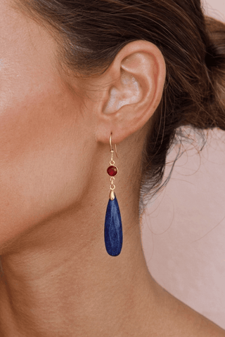 Close-up of an ear wearing a blue lapis teardrop earring with a garnet gemstone on a neutral background